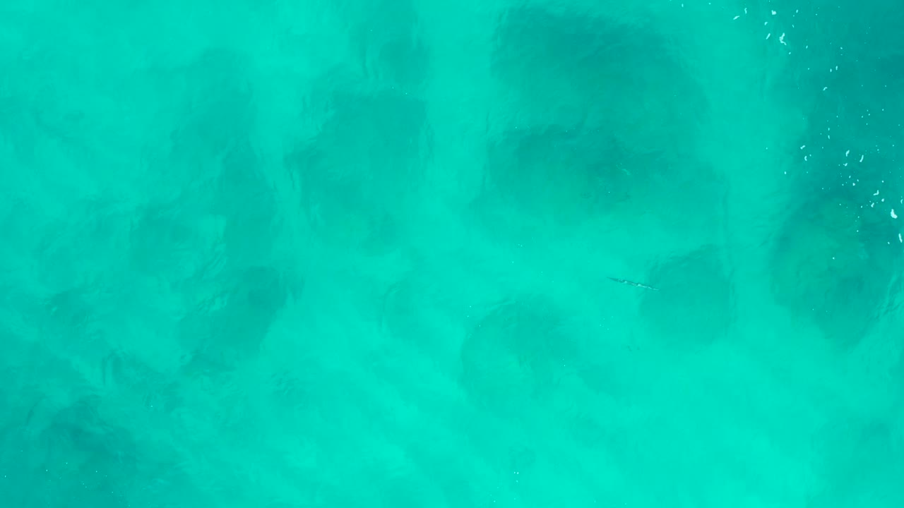 Overhead aerial frame of teal water in Baja California Sur with a clearer vertical trail of tiny bubbles and shadowy patches under the surface, no visible people.