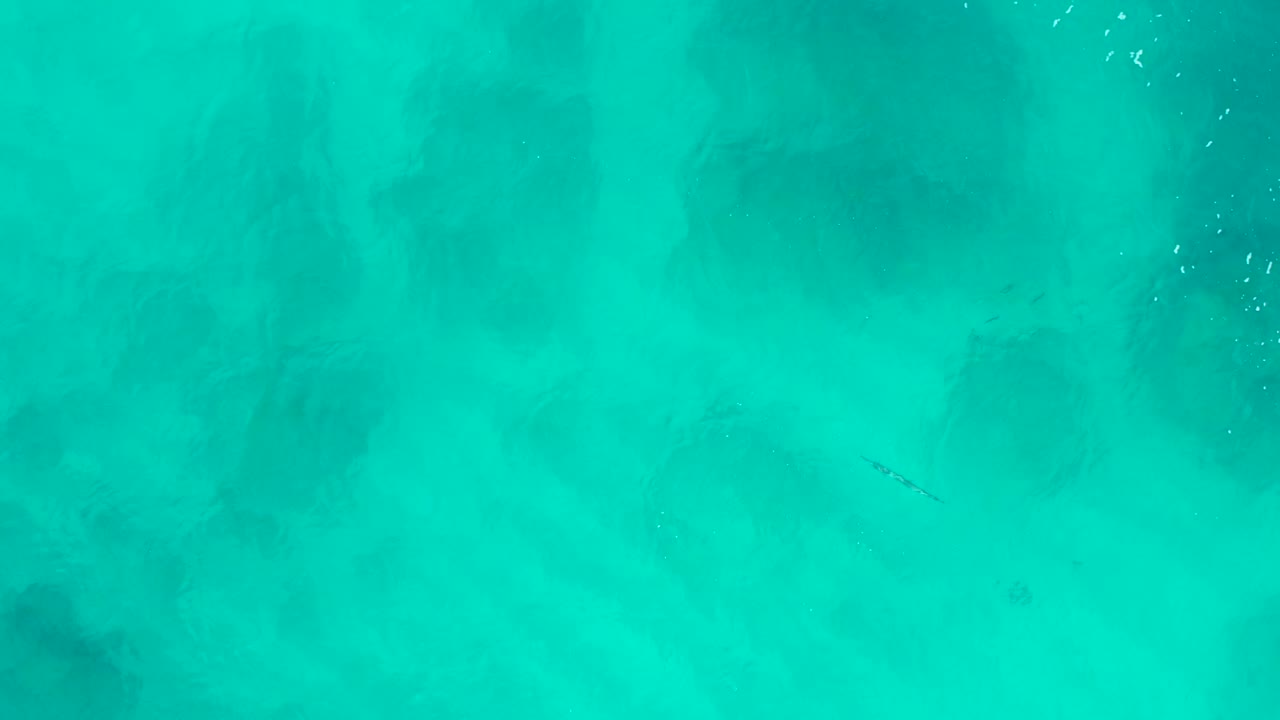 Aerial frame over turquoise water in Baja California Sur, showing darker underwater patches and small white surface glints near the upper area with no visible people.