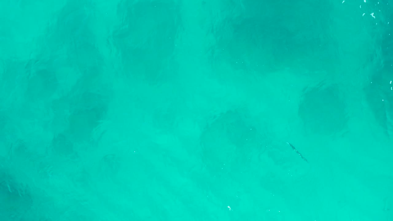 Top-down aerial frame of turquoise shallow ocean water in Baja California Sur, Mexico, with soft dark submerged patches and no visible people.