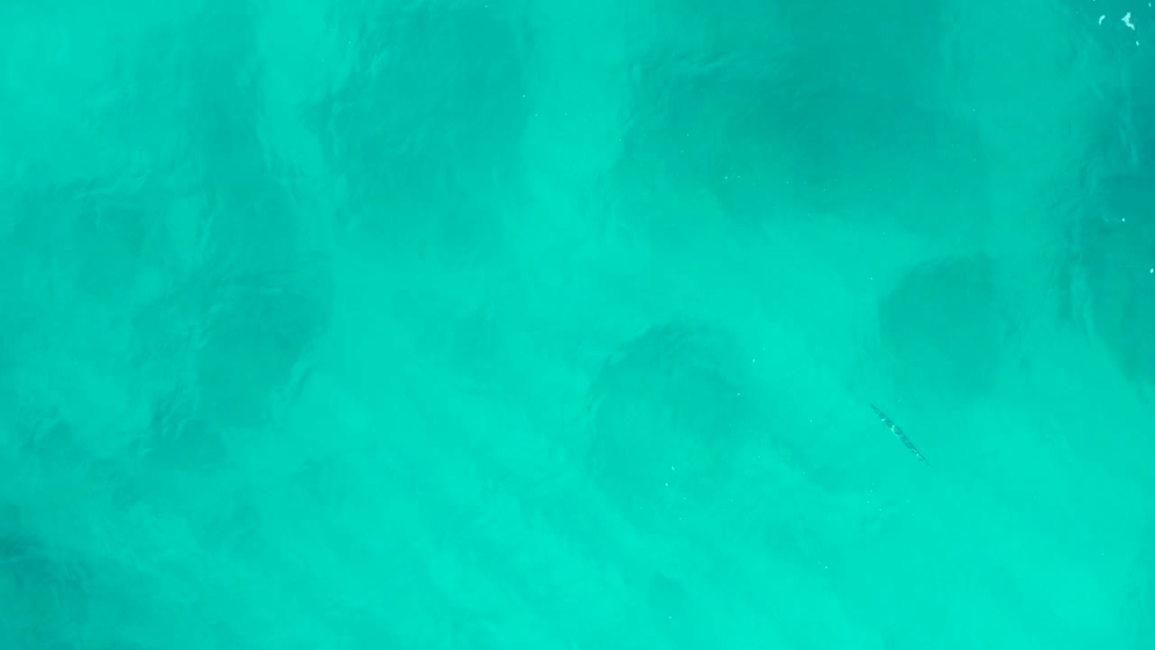 Straight-down aerial frame of turquoise shallow water in Baja California Sur, Mexico, with cloudy dark patches below the surface and no visible people.