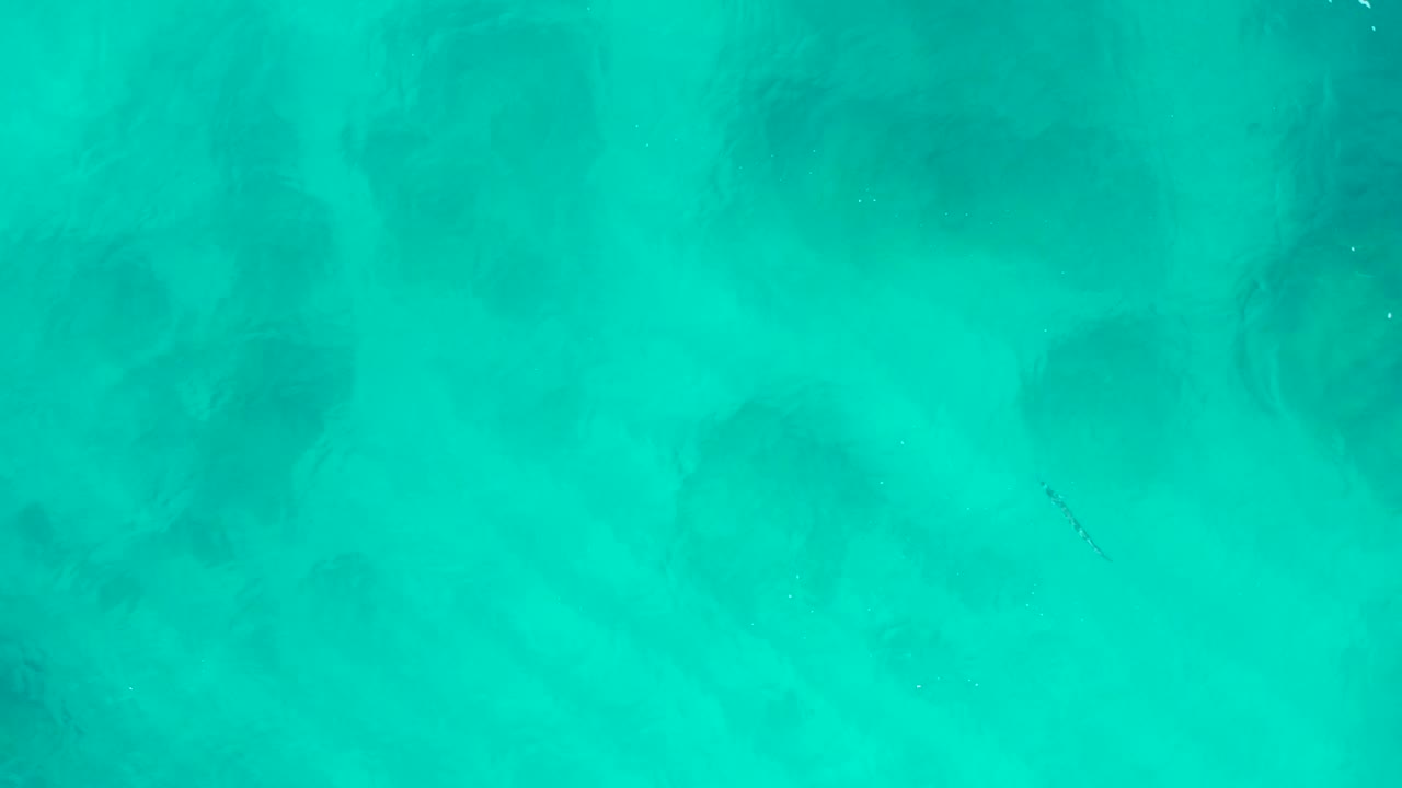 Aerial turquoise water with faint ripples and dark underwater areas.