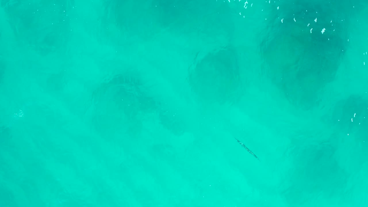 Aerial view over turquoise water in Baja California Sur with soft dark underwater shadows and no visible people.