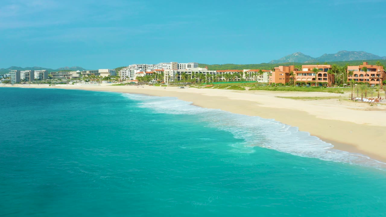 Opening aerial frame over Desert Park Natural Reserve in Baja California Sur, Mexico, with turquoise ocean at left, pale beach at right, hotels along the shore, distant mountains, and no visible people.