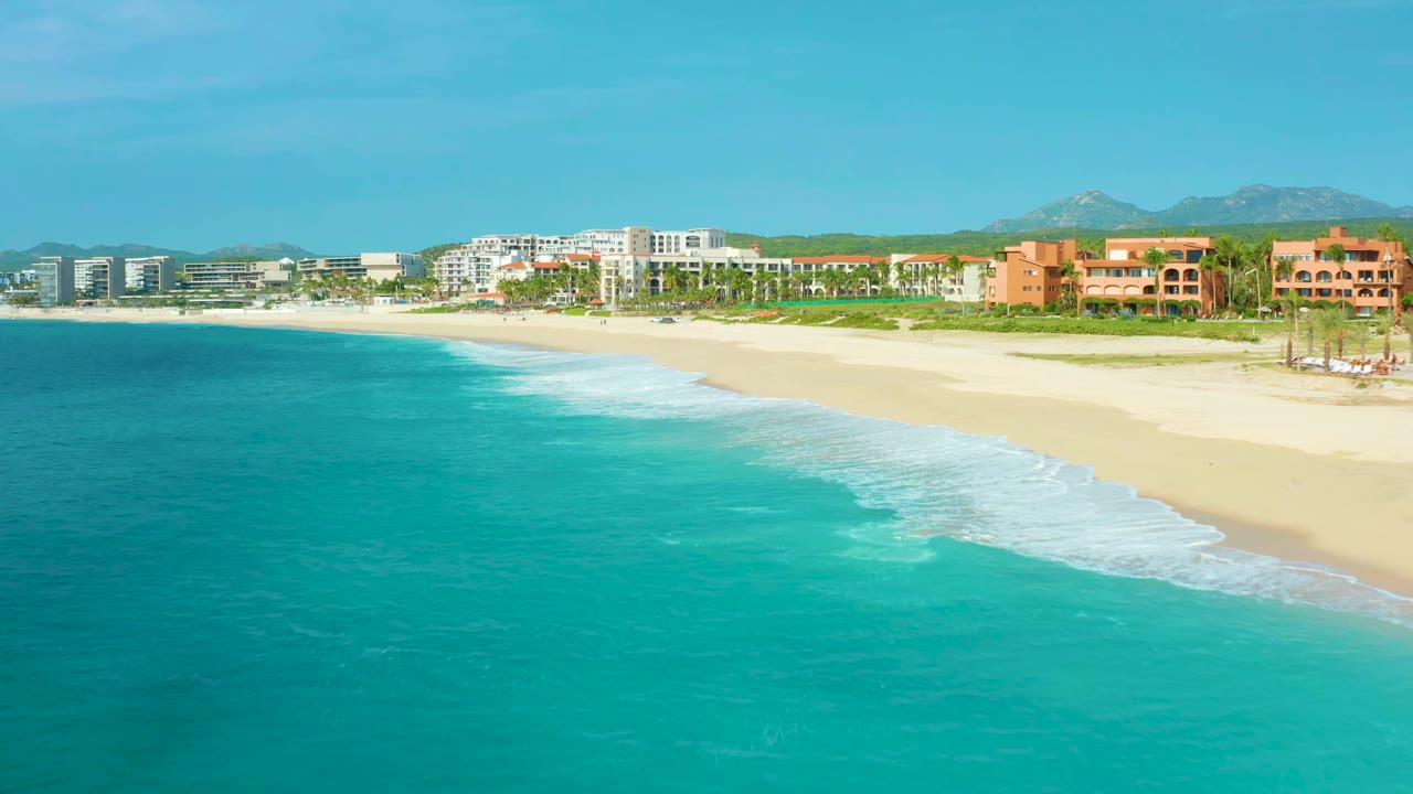 Aerial view at 0:01 over Desert Park Natural Reserve, Baja California Sur, Mexico, showing a broad band of turquoise water, surf curling onto sand, hotel buildings across the shoreline, and no visible people.