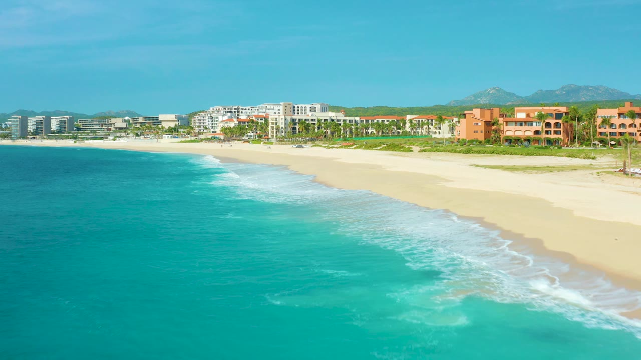 Mid-clip aerial frame over Desert Park Natural Reserve, Baja California Sur, Mexico, with clear turquoise water in front, a fresh line of foam on the sand, hotels behind coastal vegetation, and no visible people.