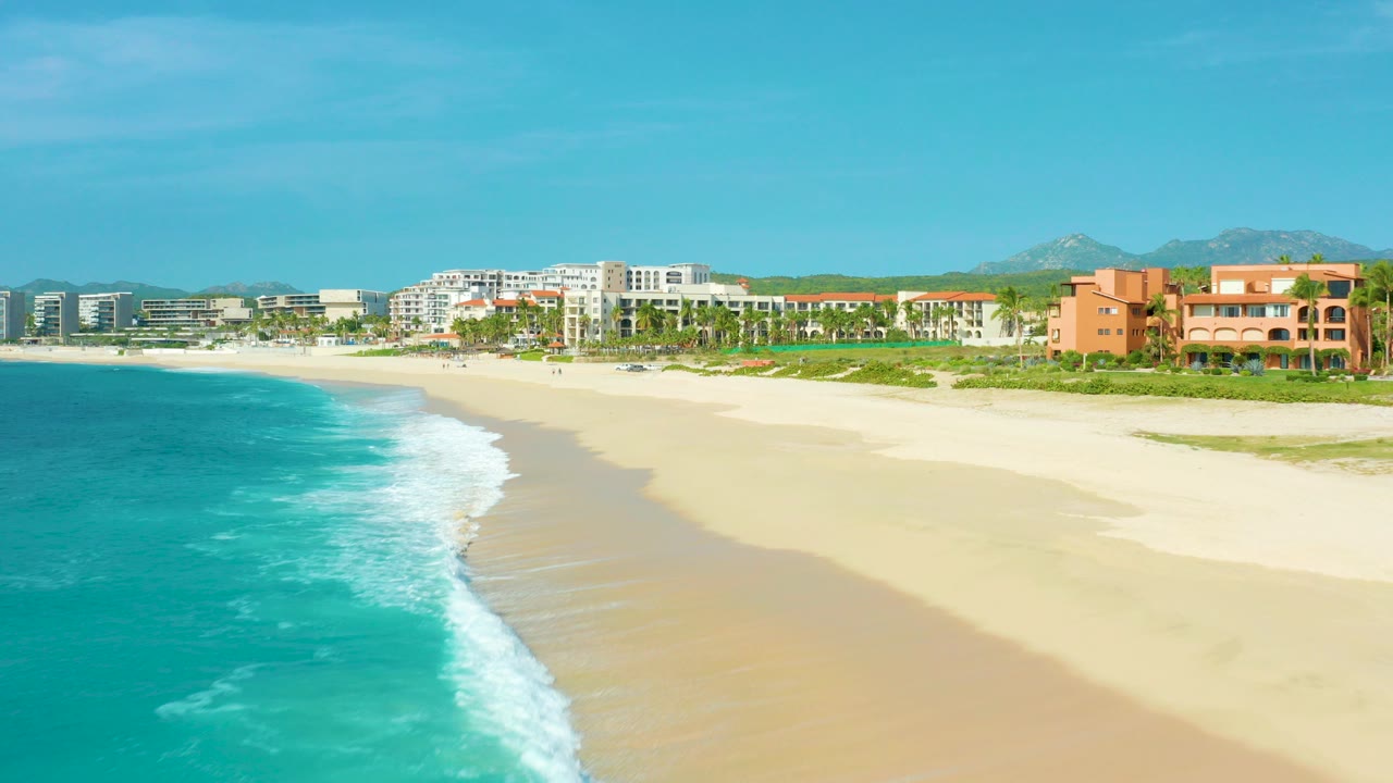 Aerial view in Desert Park Natural Reserve, Baja California Sur, with turquoise surf along the left edge, pale sand sweeping toward resort buildings, distant mountains, and no visible people.