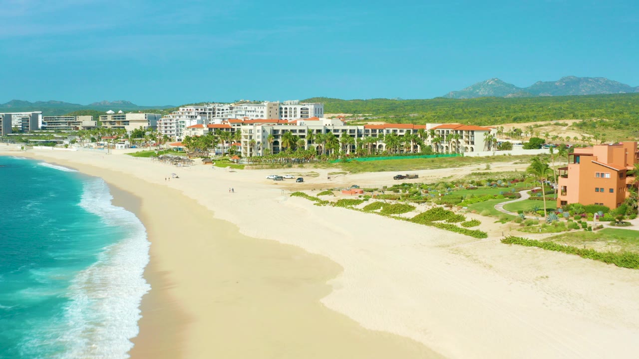 Opening aerial frame over Desert Park Natural Reserve in Baja California Sur, with turquoise water at left, pale beach below, resort buildings beyond the dunes, green hills in back, and no visible people.