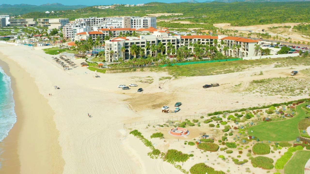 High aerial frame of a Baja California Sur beachside resort with terracotta roofs, pale sand, rows of umbrellas, scattered vehicles, ocean surf at left, and no visible people.