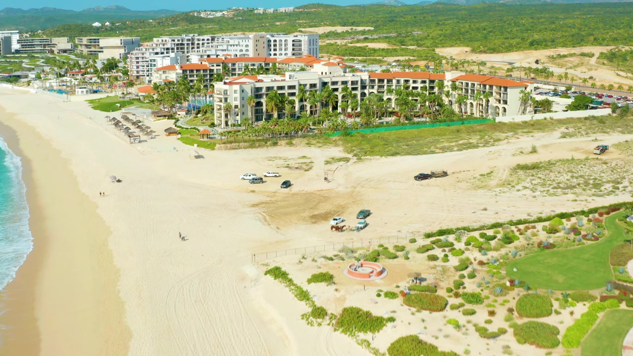 Aerial frame over the Baja California Sur shoreline showing the sandy beach foreground, ocean edge, resort buildings, terracotta roofs, umbrellas, parked vehicles, and no visible people.