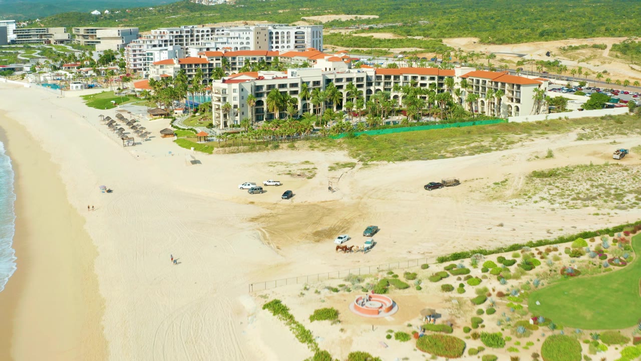 Wide drone frame of the Baja California Sur resort complex with open sand, small vehicles on the beach, planted green areas, palms, terracotta roofs, and no visible people.