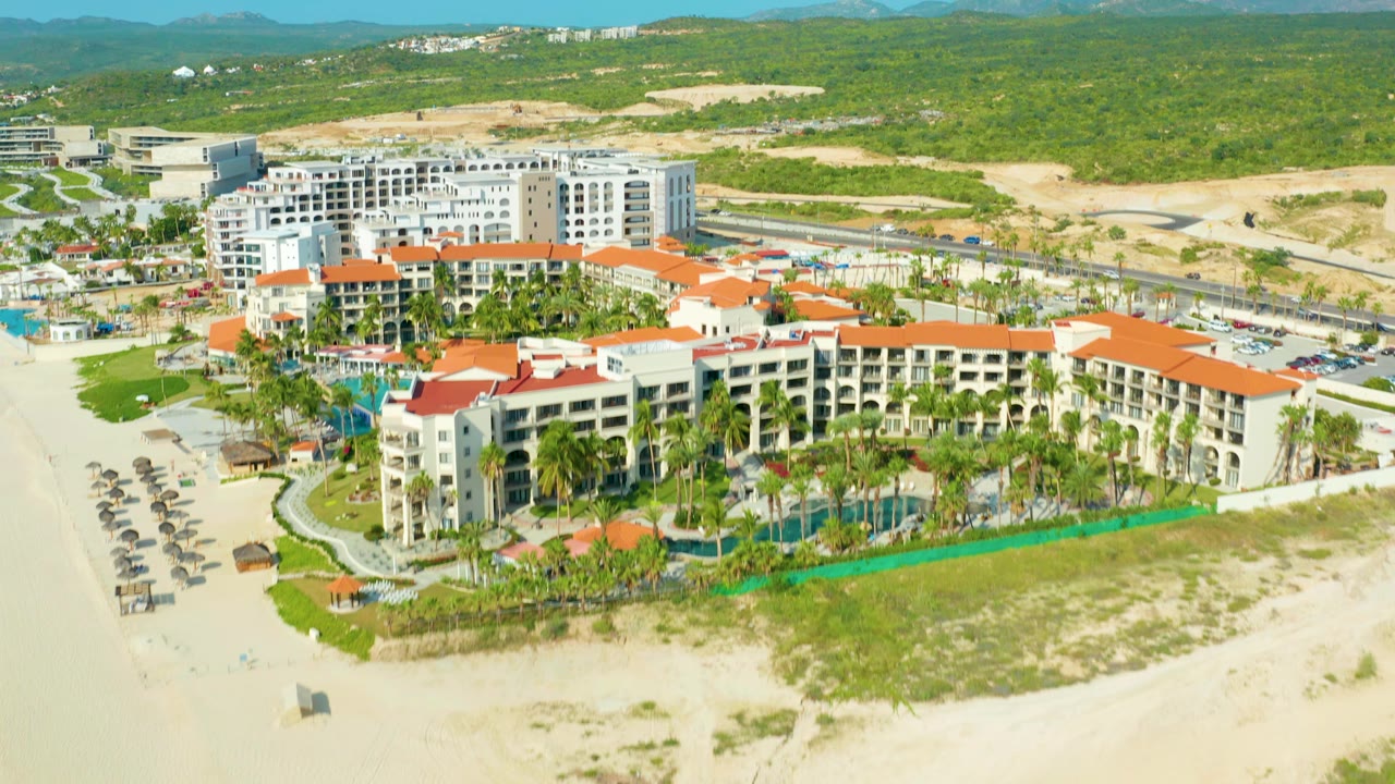 Aerial view in Baja California Sur showing the resort centered in frame, with pool courtyards and palms below, rows of beach umbrellas at left, a highway behind the buildings, and no visible people.