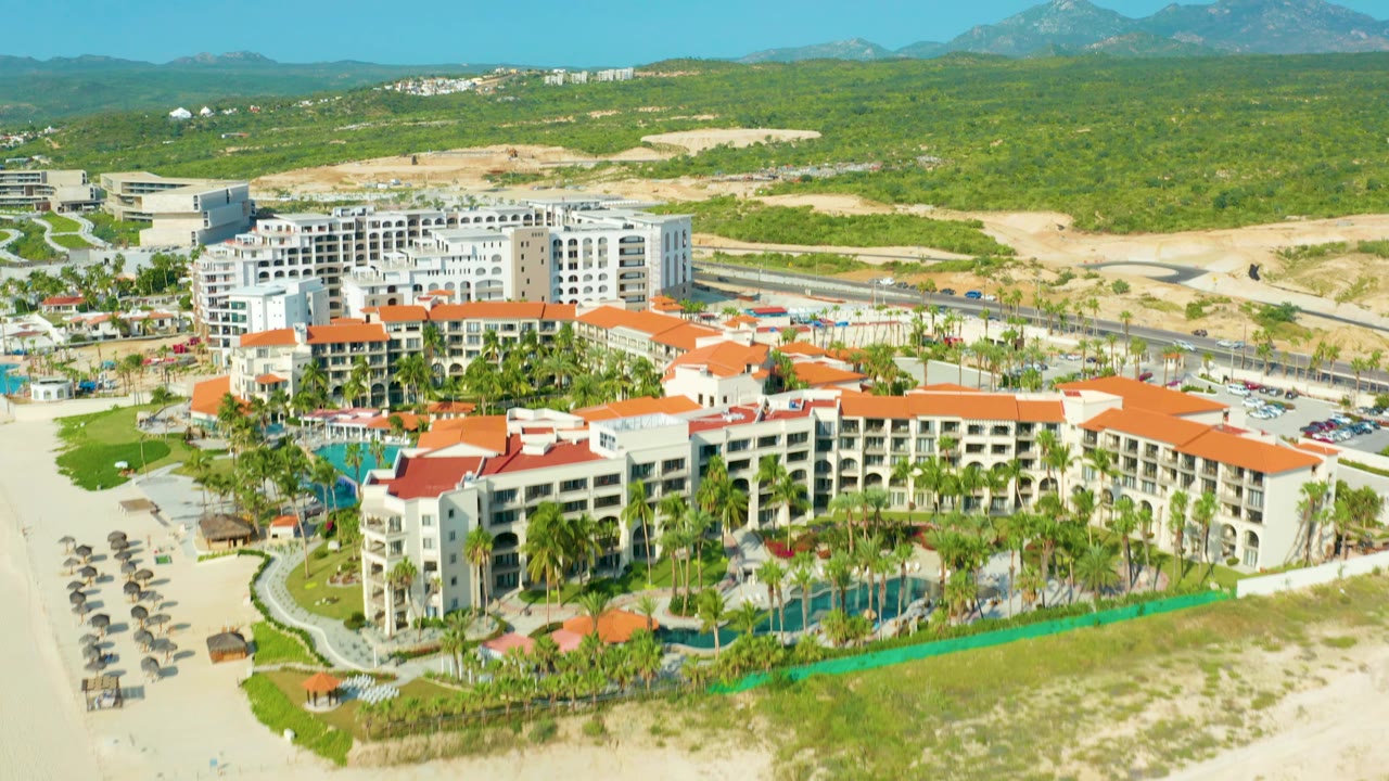 High-angle Baja California Sur resort view with more of the complex shifted to the lower right, beach umbrellas along the left sand, a highway running behind the property, desert scrubland, mountains, and no visible people.