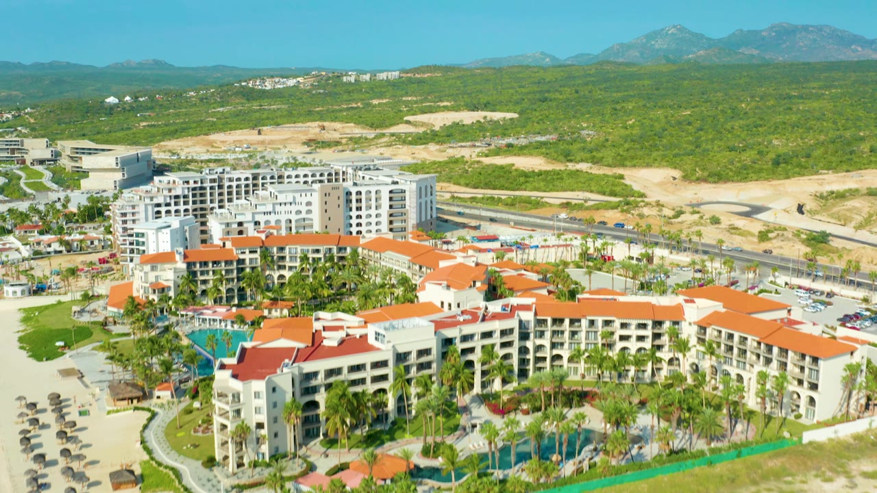 Baja California Sur aerial frame showing the resort buildings across the lower half, pools and palm-lined grounds in front, a highway and sandy open lots behind, distant mountains, and no visible people.