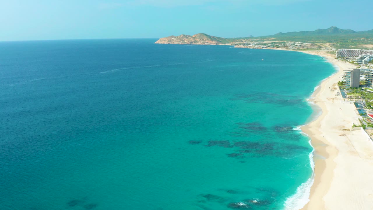 High aerial view over Desert Park Natural Reserve in Baja California Sur, Mexico, with turquoise shallows, deep blue ocean, pale beach, distant headland, resort buildings at the right edge, and no visible people.