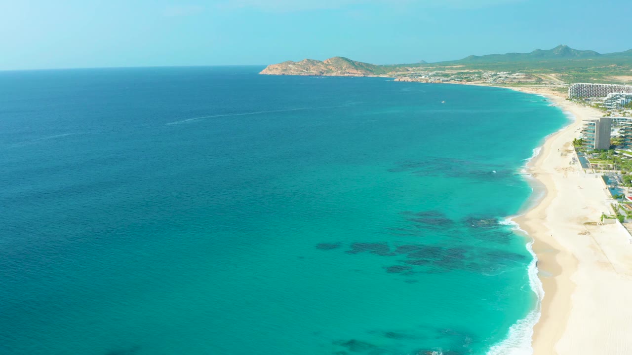 Wide drone composition in Desert Park Natural Reserve showing a sweeping beach, bright turquoise water, darker reef or rock shapes below the surface, resort buildings along the right margin, and no visible people.
