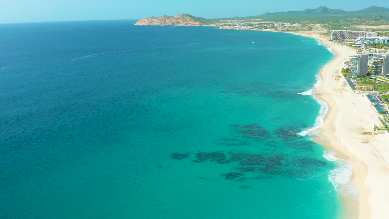 Aerial frame over Desert Park Natural Reserve in Baja California Sur showing turquoise shallows, dark underwater patches, pale sand, resort buildings on the right edge, distant hills, and no visible people.