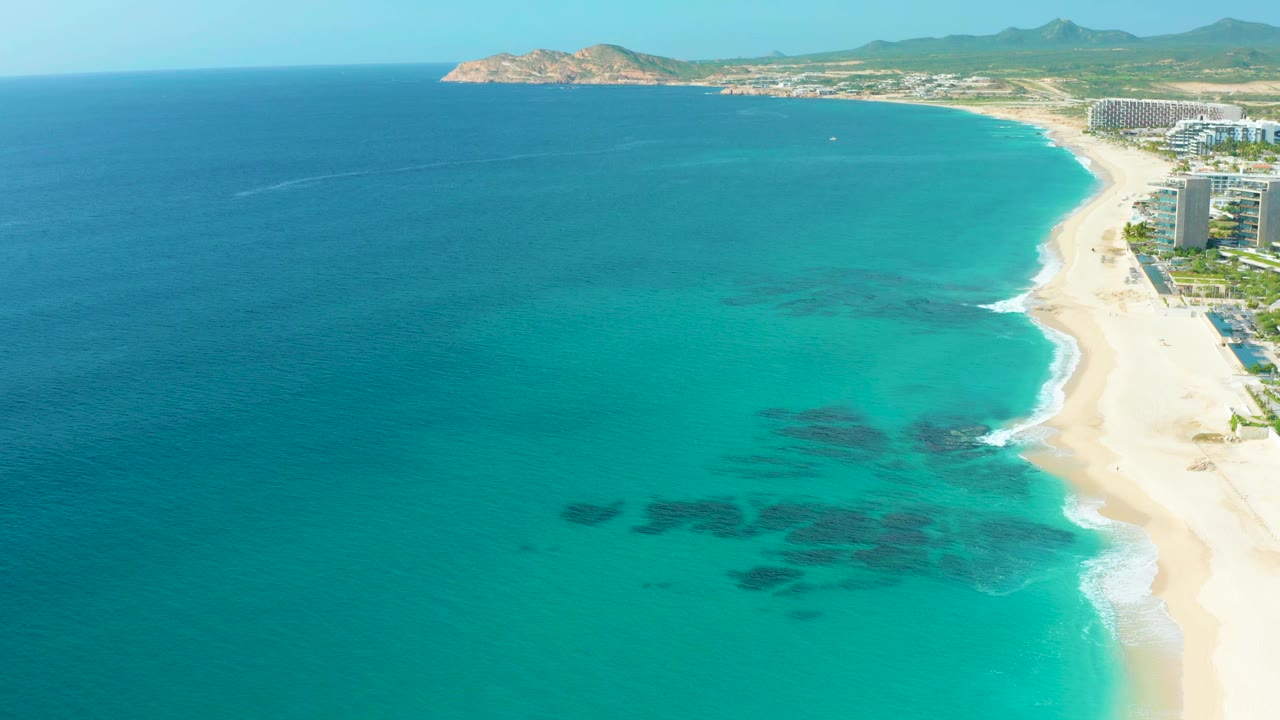 Second aerial frame in Baja California Sur with more open turquoise water in the center, reef-like dark shapes below the surface, the curved beach on the right, resort towers, and no visible people.