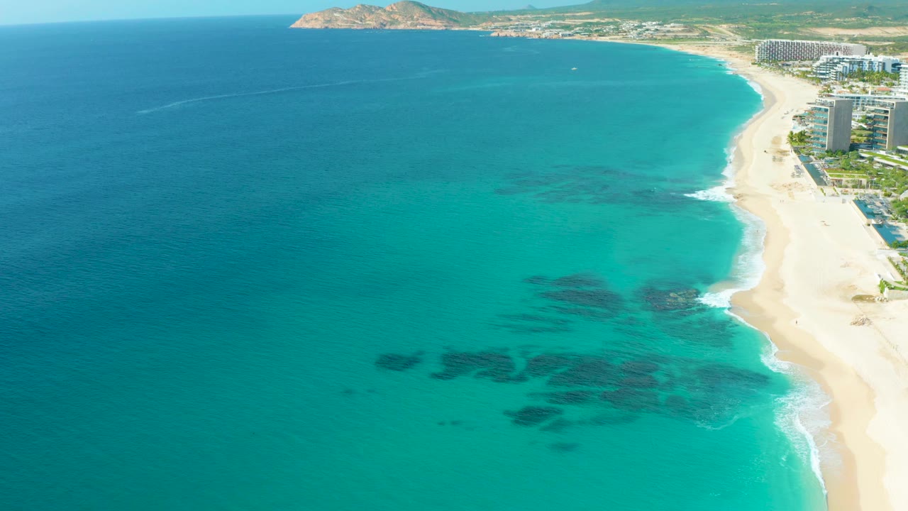 Mid-clip aerial frame in Baja California Sur showing the sandy beach running down the right side, white surf curling at the edge, multiple dark underwater areas in turquoise water, resort structures, and no visible people.