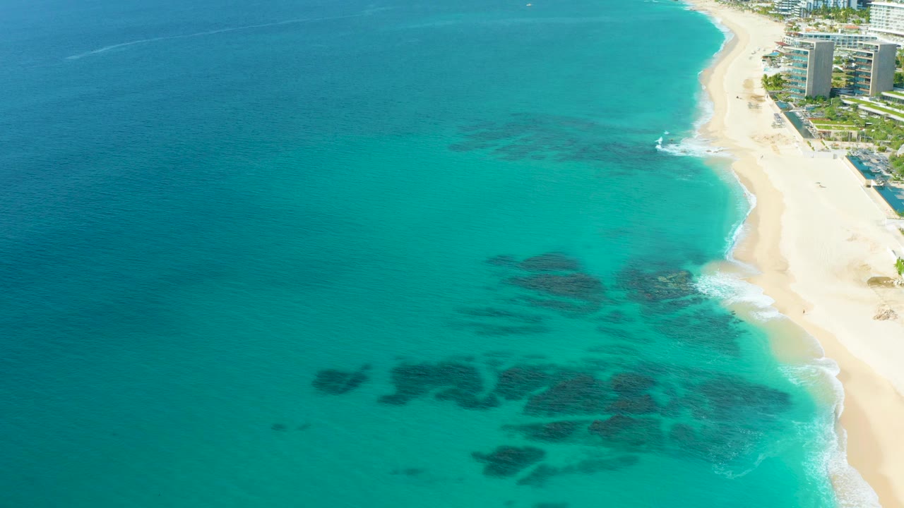 Aerial frame over Baja California Sur coastline with open turquoise water dominating the view, dark submerged formations near the lower center, pale beach along the right edge, and no visible people.