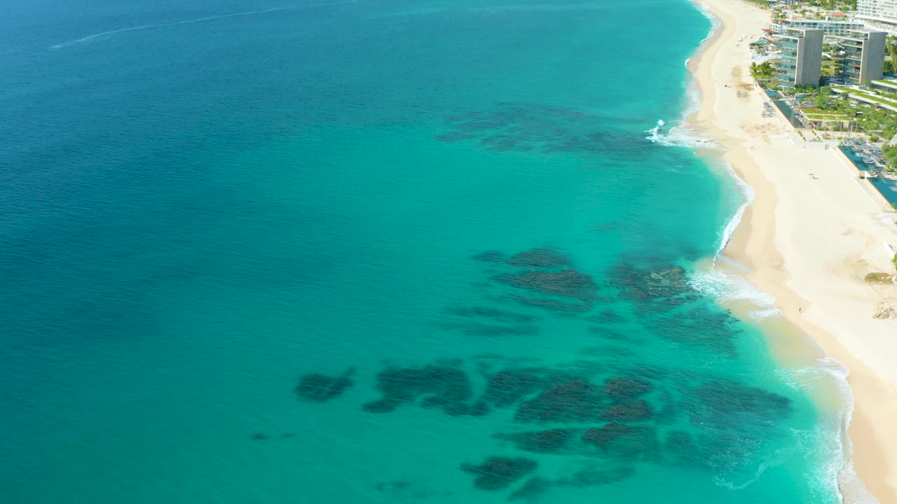 High aerial view in Baja California Sur with dark reef-like patches visible through turquoise water, a bright sandy beach curving along the right side, shoreline buildings at the top right, and no visible people.