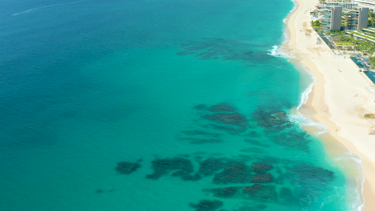 Baja California Sur aerial frame with a broad field of turquoise water, darker submerged patches lower in frame, a narrow surf line on the right, beachfront buildings near the top edge, and no visible people.