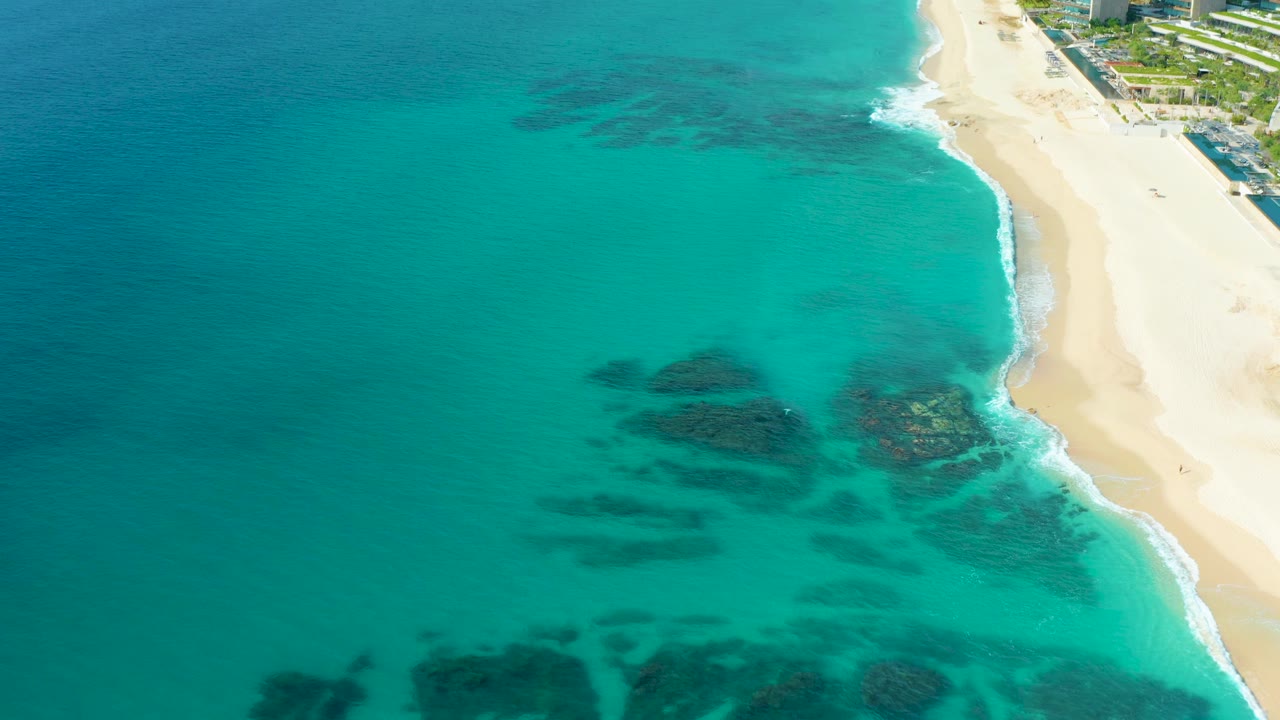 Aerial frame over the Baja California Sur coast with open turquoise water across the left, the sand beach along the right edge, dark reef forms under the surface, and no visible people.
