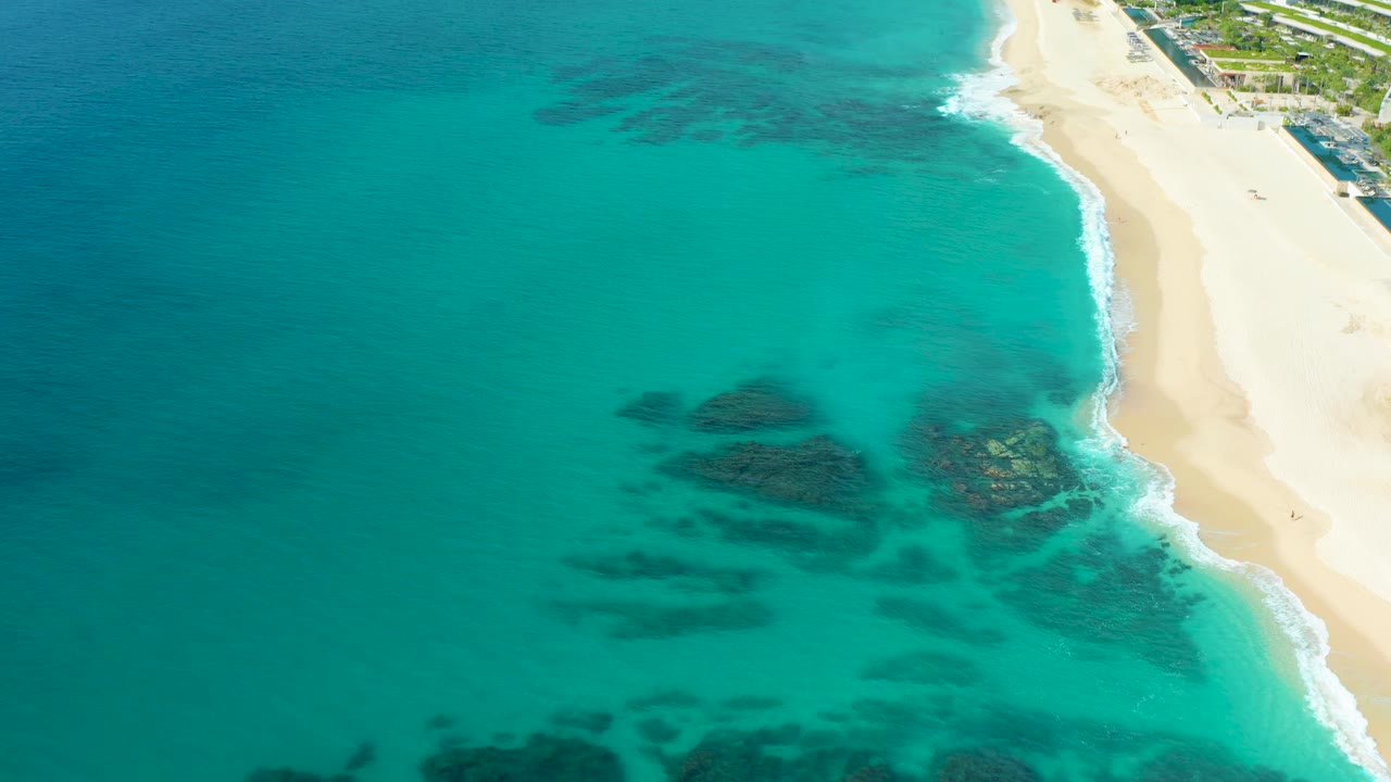 Aerial view of Desert Park Natural Reserve coastline where the beach curves down the right side, white surf traces the edge, and dark reef patches show through the turquoise water with no visible people.