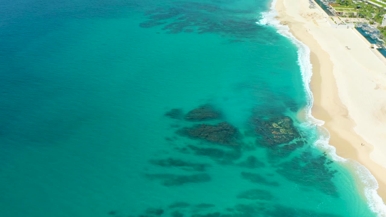 Drone view of clear turquoise water, reef patches, surf, and sandy shoreline.