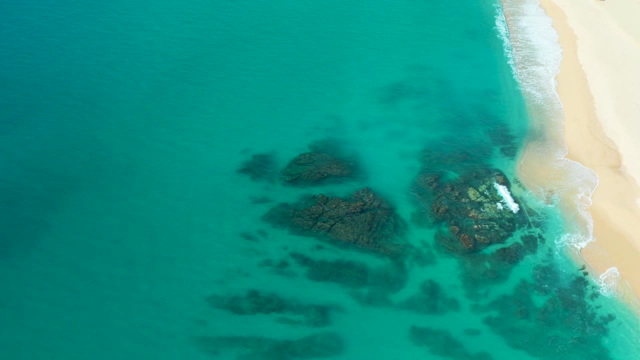 Overhead drone frame from Desert Park Natural Reserve in Baja California Sur showing turquoise water around dark rocks beside a pale beach, with no visible people.