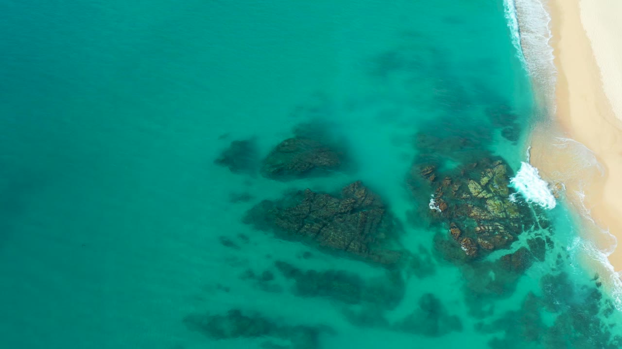 Overhead frame showing the rock group slightly lower in the composition, with turquoise water surrounding it and the sandy Baja California Sur shore on the right, with no visible people.