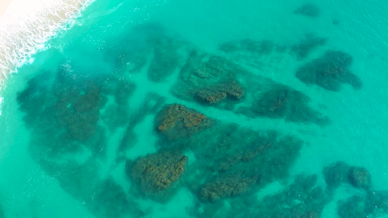 Opening aerial frame over Desert Park Natural Reserve, Baja California Sur, showing turquoise shallow water, dark reef patches below the surface, white surf at the upper left, and no visible people.