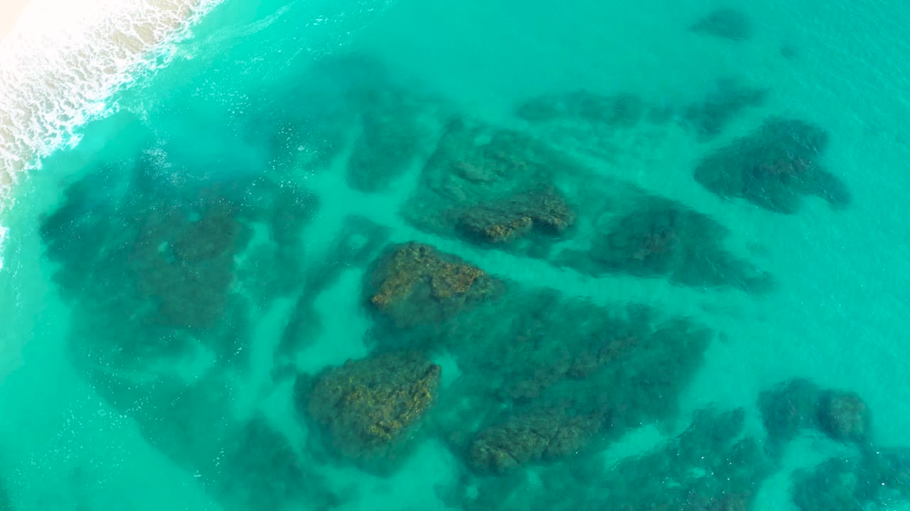 Aerial view one second in over Desert Park Natural Reserve, Baja California Sur, with the pale beach edge still visible at upper left and clustered dark reef shapes under aquamarine water, with no visible people.