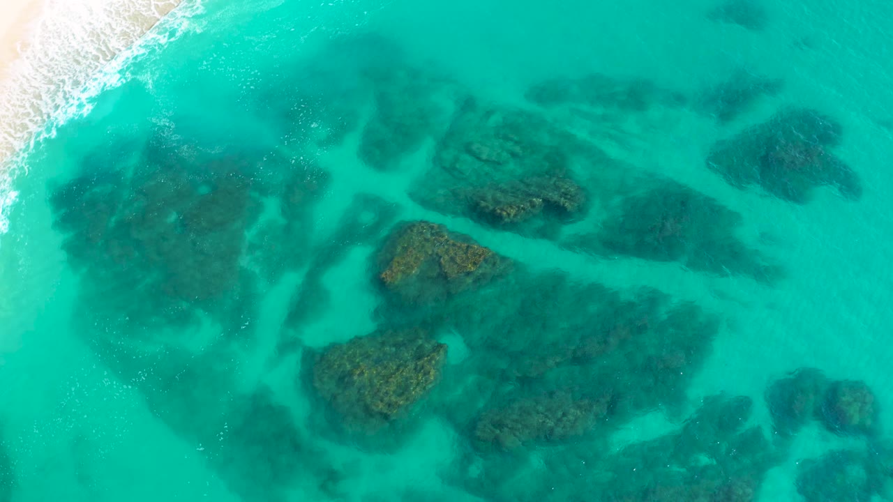 Aerial frame over Desert Park Natural Reserve, Baja California Sur, showing a wider spread of dark underwater reef patches across turquoise water, a thin white shore break at upper left, and no visible people.