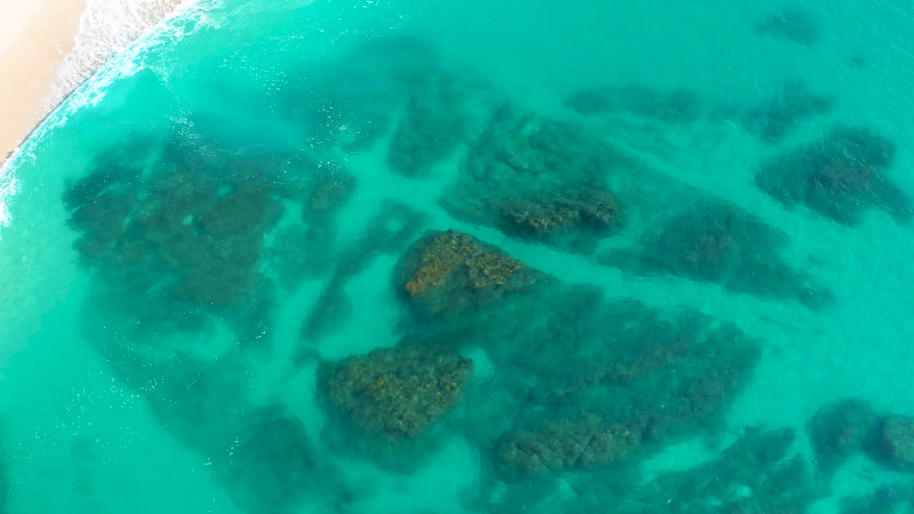 Mid-clip aerial view at Desert Park Natural Reserve, Baja California Sur, where turquoise lagoon water covers dark underwater rocks and the shoreline foam runs diagonally near the upper left, with no visible people.