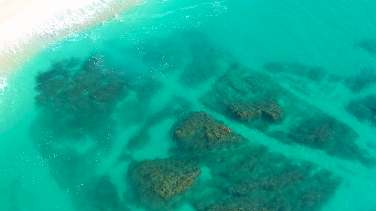 Aerial view in Desert Park Natural Reserve, Baja California Sur, with the surf line still at the top left and several dark rock masses visible through bright turquoise water, with no visible people.