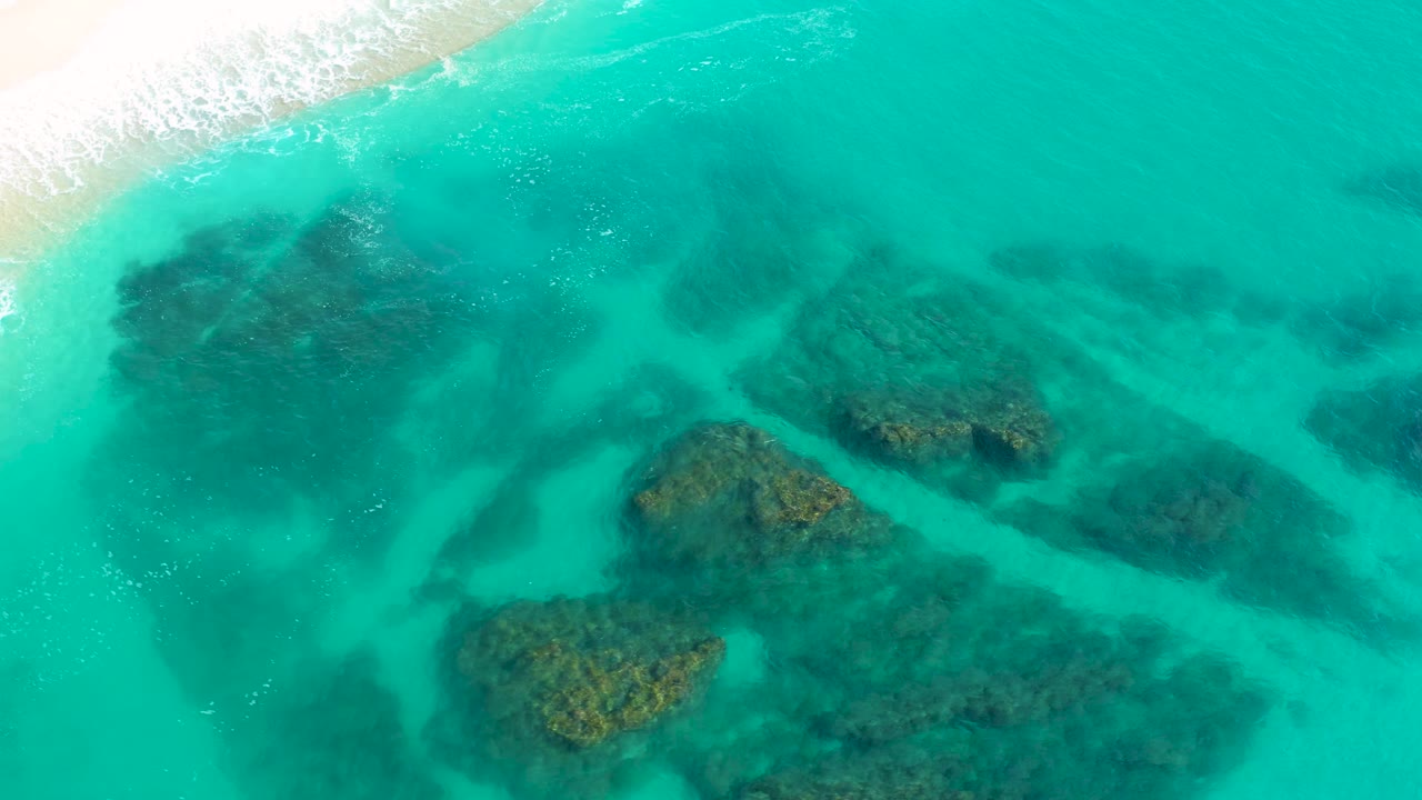 High-angle drone frame over Desert Park Natural Reserve, Baja California Sur, showing larger submerged rock patches under teal water with a narrow strip of foamy beach surf at the upper left and no visible people.