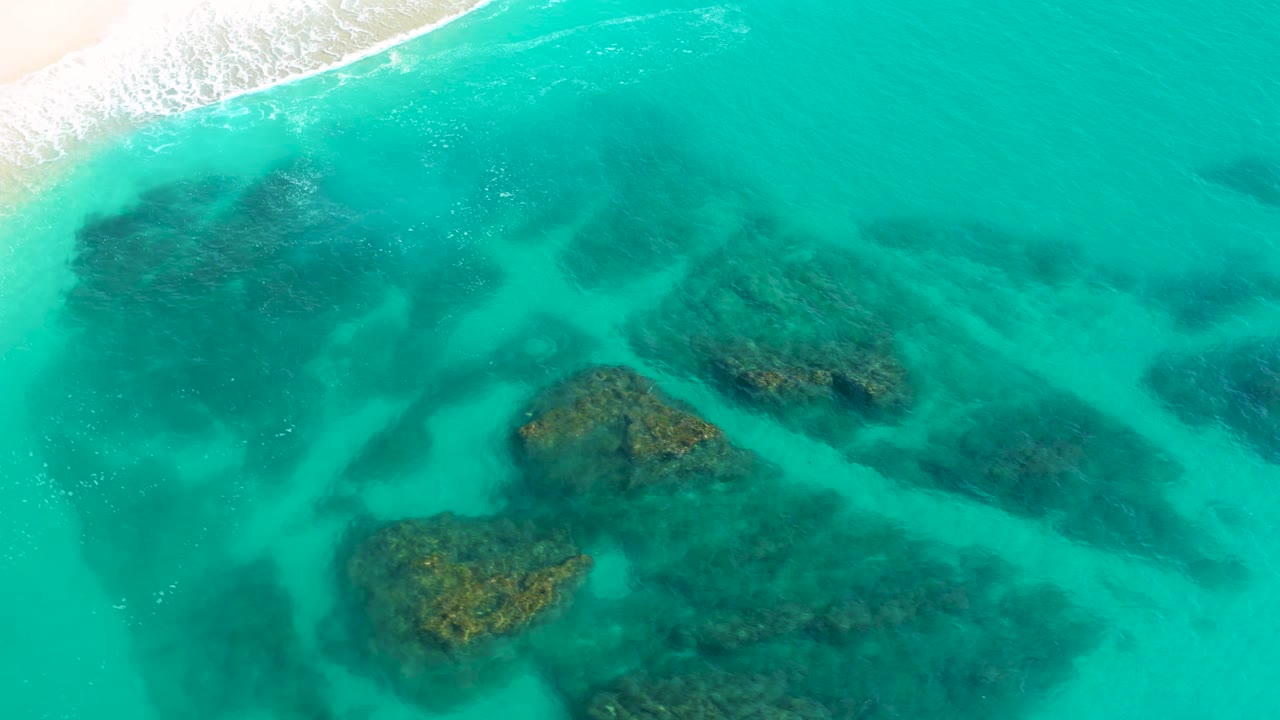 Drone view from Desert Park Natural Reserve, Baja California Sur, with the beach and white wash pushed toward the upper left while broad submerged rock shapes spread through the turquoise shallows, with no visible people.