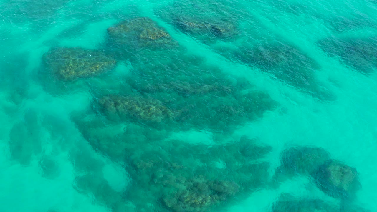 Top-down aerial frame over turquoise water in Desert Park Natural Reserve, Baja California Sur, with dark reef patches below the surface and no visible people.