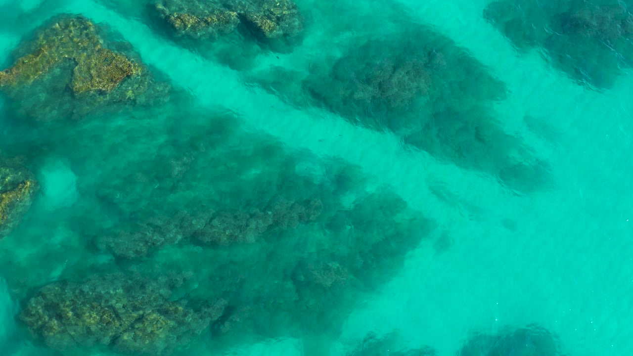 Top-down aerial frame shows a pale diagonal sandy channel cutting between darker submerged rocks in Desert Park Natural Reserve, Baja California Sur, with no visible people.