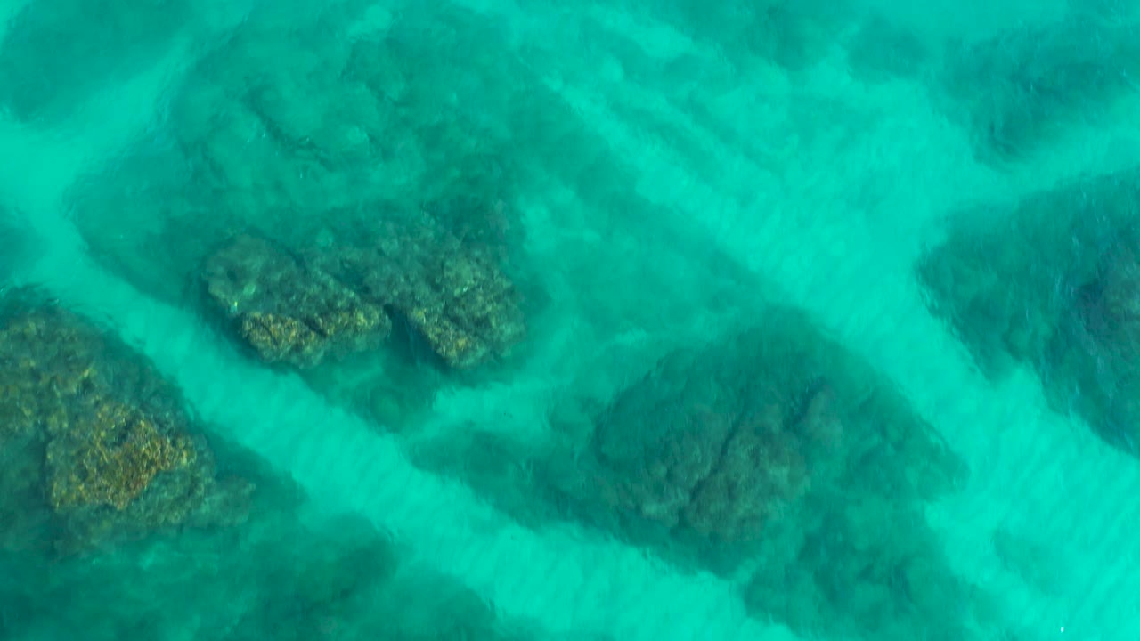 Overhead view of shallow turquoise water with clustered dark reef rocks and lighter sandy bottom visible in Desert Park Natural Reserve, Baja California Sur, Mexico, with no visible people.