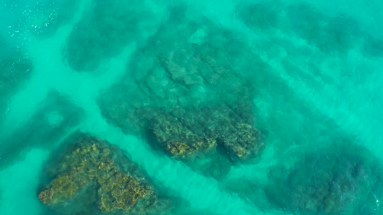 Top-down Baja California Sur water view with dark green-brown rocks under turquoise water, faint surface highlights near the upper edge, and no visible people.