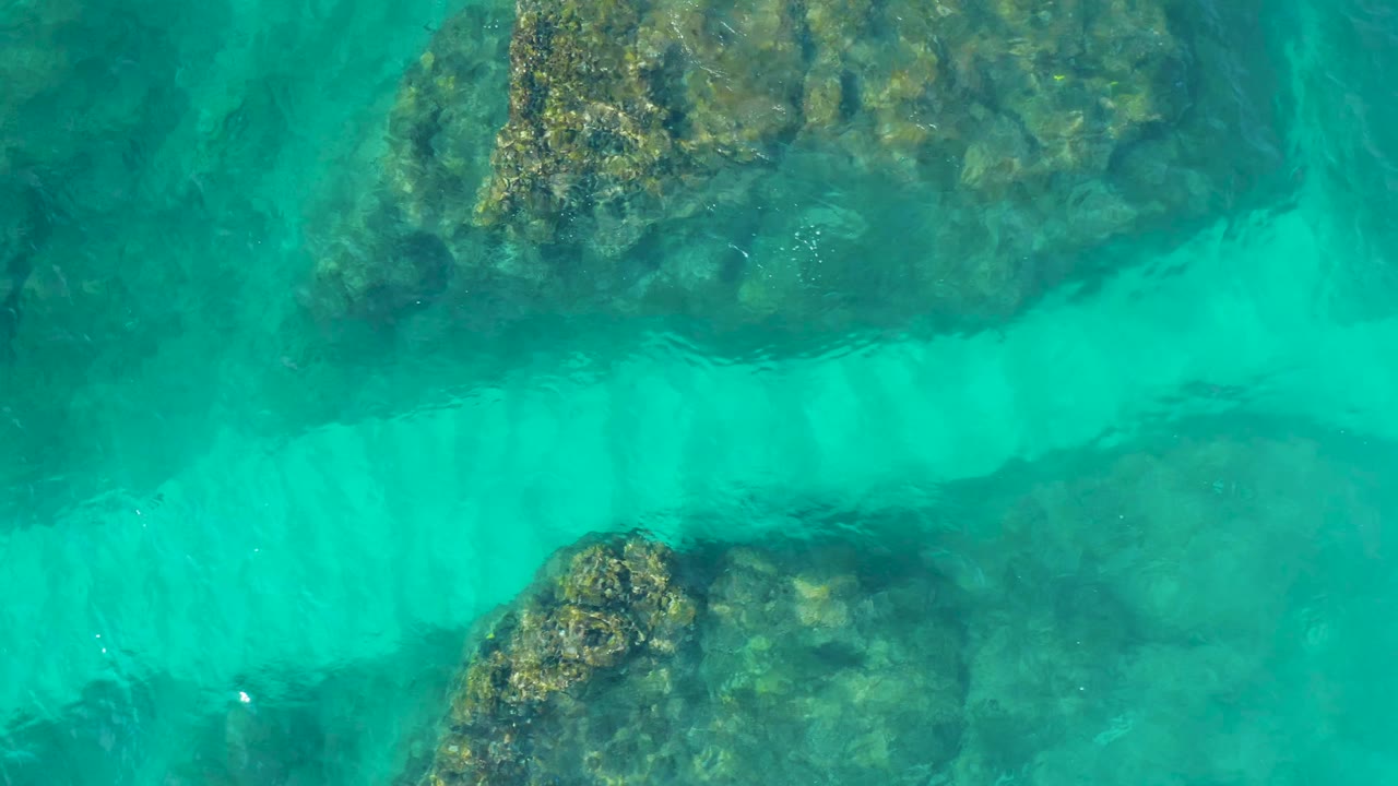 Overhead aerial view in Baja California Sur with a broad turquoise channel crossing between mottled brown-green rock ledges and no visible people.