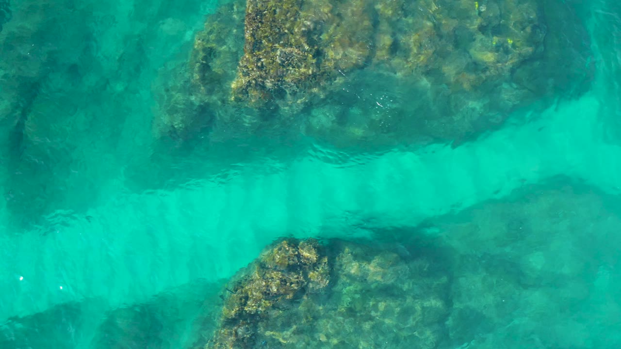 Top-down frame over the Mexico reserve showing translucent blue-green water, a pale underwater band, and large textured rocks along the upper and lower edges with no visible people.