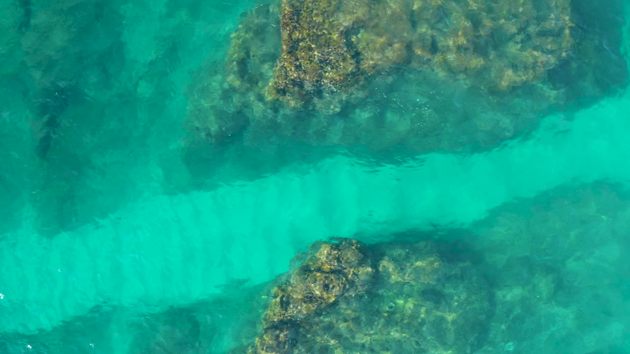 Aerial top-down view of Desert Park Natural Reserve water with a large mottled rock formation near the upper frame and a turquoise channel below, with no visible people.