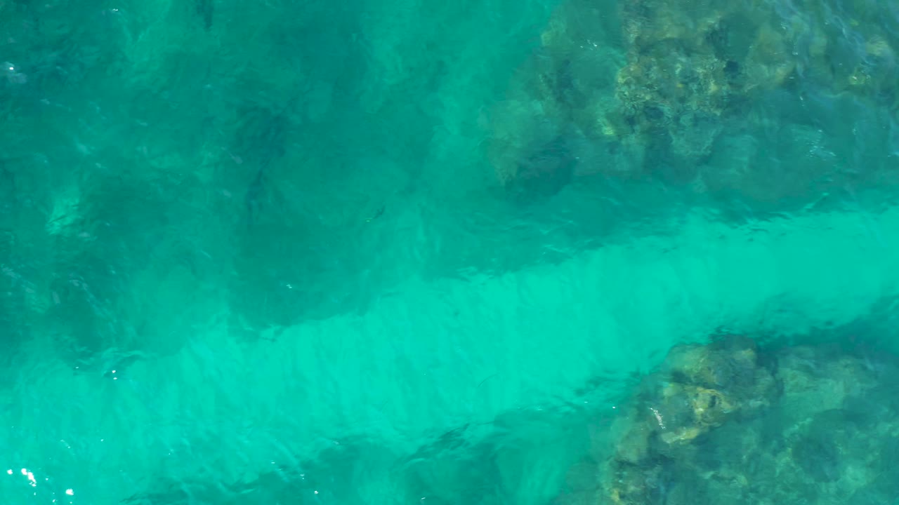 Clear teal water fills the frame over a broad dark reef band and pale sandy shallows in Baja California Sur, Mexico, with no visible people.