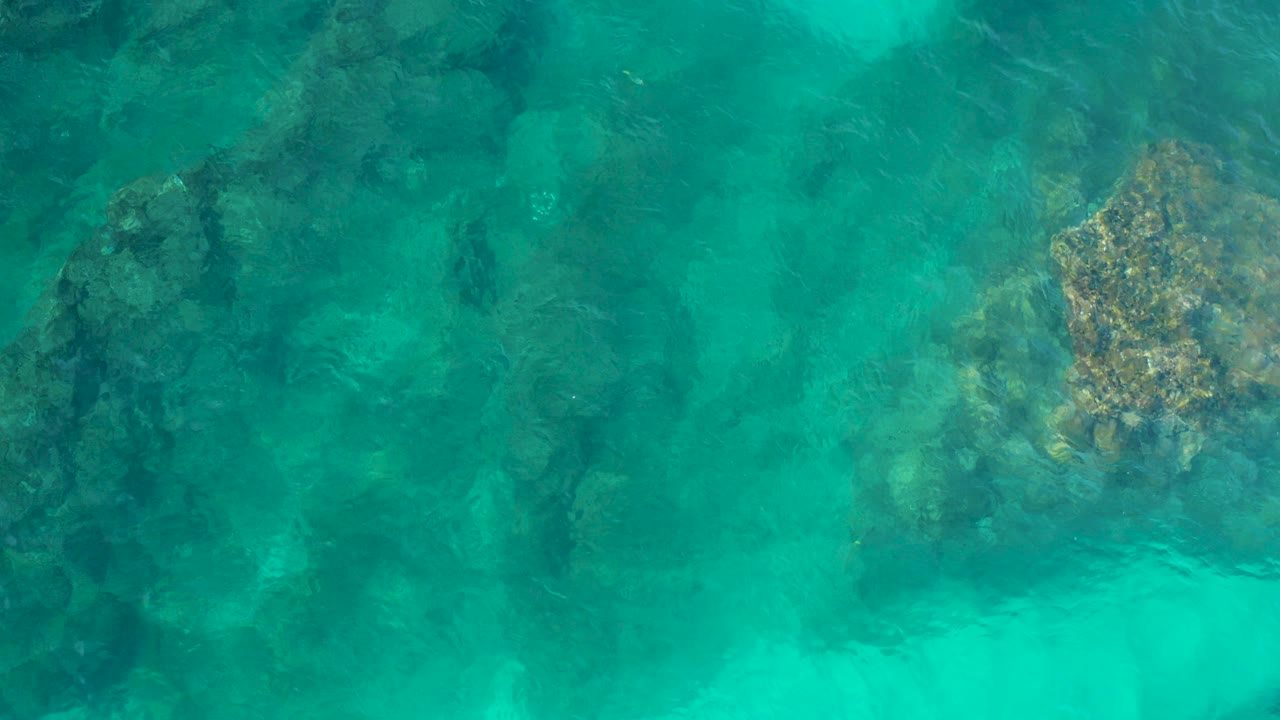 Top-down aerial view of turquoise water over dark submerged rocks and lighter seabed patches in Desert Park Natural Reserve, Baja California Sur, with no visible people.