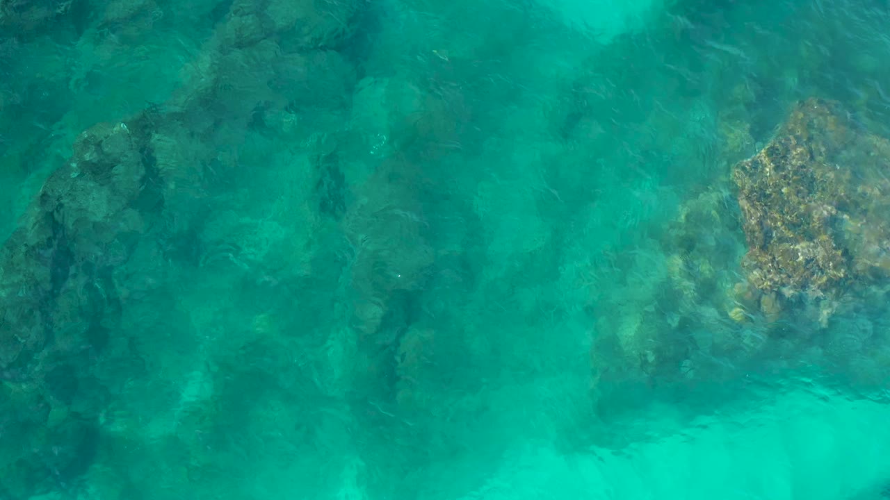 Overhead frame of clear teal water in Desert Park Natural Reserve, Baja California Sur, with a larger tan-brown submerged rock cluster to the right and no visible people.