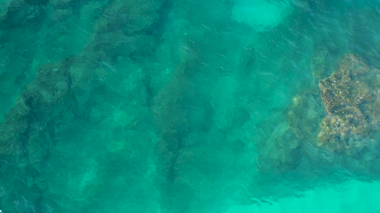 Top-down view of rippled turquoise water over scattered dark rocks and pale green seabed areas in Desert Park Natural Reserve, Baja California Sur, with no visible people.