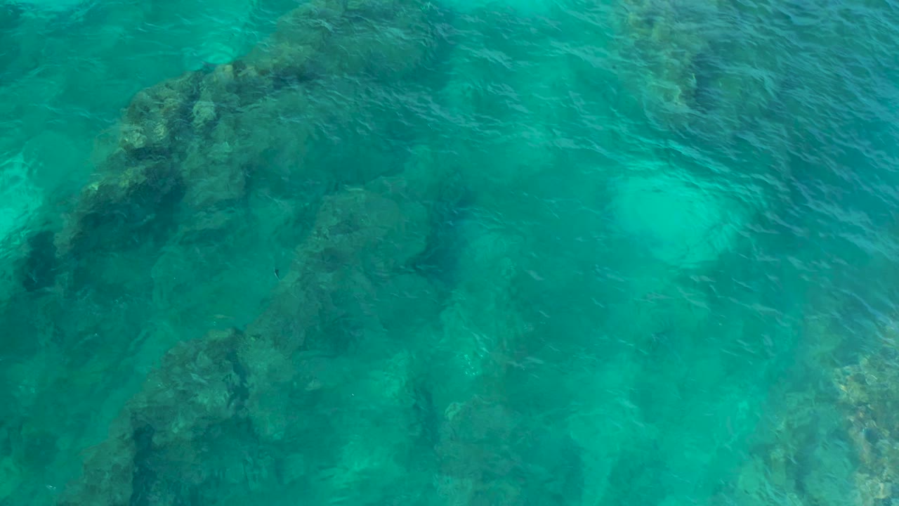 Aerial frame of clear shallow water with a broad dark rock formation near the upper center and lighter sand patches visible below, with no visible people.