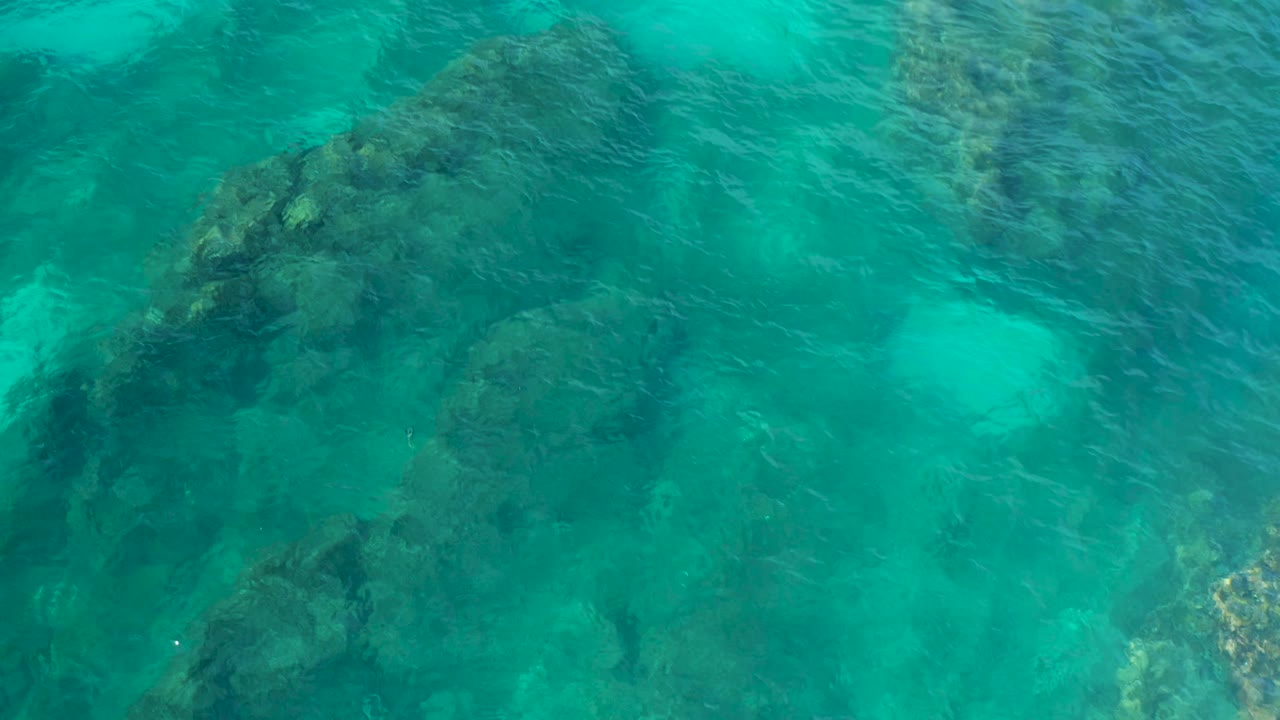 Top-down view in Baja California Sur showing teal ocean surface, darker submerged rock shapes, and small pale areas of seafloor, with no visible people.